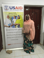 WI-HER'S Kebbi State Gender, Social Inclusion and Community Engagement Advisor, Nafisa Zaki, poses next to the USAID Integrated Health Program Banner.